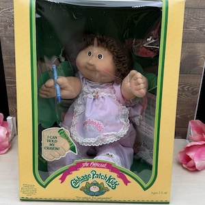 May include: A Cabbage Patch Kids doll with brown yarn hair, wearing a light purple dress with a white lace-trimmed apron. The doll holds a blue crayon in its hand. The packaging features the text I CAN HOLD MY CRAYON and The Official Cabbage Patch Kids.
