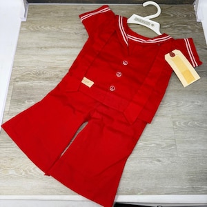 May include: A bright red sailor-style outfit featuring a short-sleeved top with a wide collar, white striped trim, and three white buttons down the front. The matching red pants have a wide-leg cut. A white plastic hanger and a beige tag are attached.