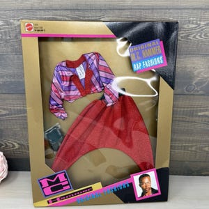 May include: A Mattel M.C. Hammer Rap Fashions doll clothing set in a gold and black box. The set includes a long-sleeved jacket with purple, pink, and red stripes and a red glittery pair of harem pants. The packaging features the text Original M.C. Hammer Rap Fashions and a small photograph of M.C. Hammer.
