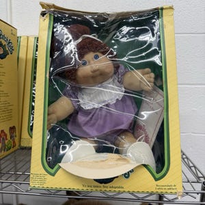 May include: A Cabbage Patch Kids doll with brown yarn hair in a ponytail, blue eyes, and a light complexion. The doll wears a purple dress with a white ruffled collar and white shoes. It is packaged in a yellow box with clear plastic.