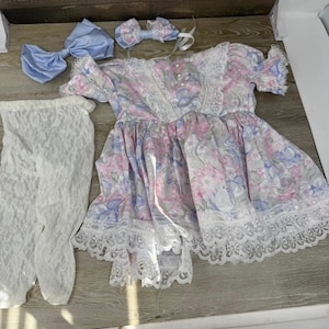 May include: A short-sleeved dress with a floral pattern in shades of pink, blue, and white, featuring white lace trim, pearl-like buttons, a matching light blue hair bow, a smaller patterned hair bow, and a pair of white lace tights.
