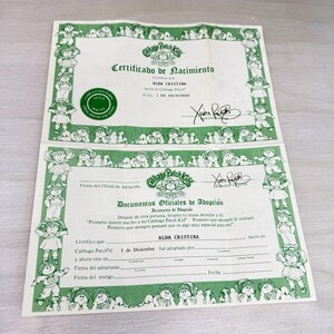 May include: A Cabbage Patch Kids birth certificate and adoption document set printed on white paper with green borders featuring illustrations of dolls. The text includes Certificado de Nacimiento, Alda Cristina, and Documentos Oficiales de Adopción.