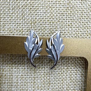 May include: A pair of silver-toned leaf-shaped earrings. Each earring features a detailed leaf design with a curved, slender stem. The leaves have a textured appearance, and the earrings are set against a neutral background.