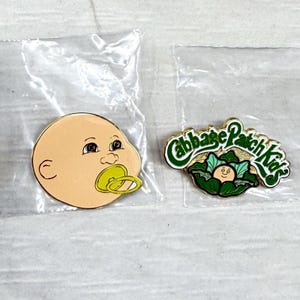 May include: Two enamel pins in clear plastic packaging. One pin is a round, peach-colored baby face with a yellow pacifier. The second pin features the green text Cabbage Patch Kids above a small illustration of a baby face inside a green cabbage.