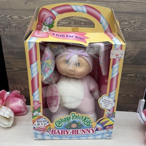 May include: A Cabbage Patch Kids Baby Bunny doll in a pink and white bunny suit with long ears, wearing a light purple bow on its head. The doll is inside a yellow cardboard box with the text A Gift For You and Cabbage Patch Kids Baby Bunny.