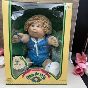 May include: A Cabbage Patch Kids doll with blonde yarn hair and blue eyes, wearing a blue sailor-style romper with a white collar. The doll holds a purple crayon in its right hand. The packaging features the text I CAN HOLD MY CRAYON!.