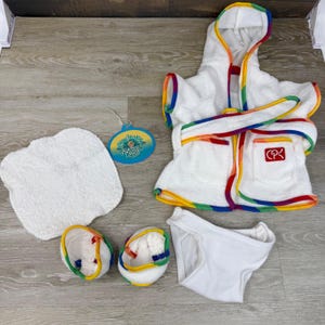 May include: A white terry cloth bathrobe with a hood and rainbow trim, a matching pair of slippers, a white washcloth, and white underwear. A blue circular tag with the text Splashin Kids is attached. A red CPK logo is on the bathrobe pocket.