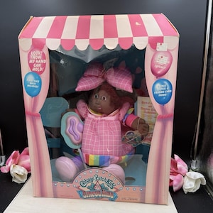 May include: A Cabbage Patch Kids Circus Kids doll with dark skin and brown hair, wearing a pink plaid dress with a matching bow. The doll is inside a pink and white striped box featuring text including Look! Now my hand can hold! and Circus Kids.