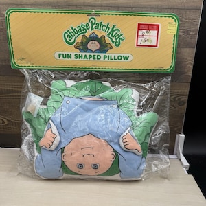 May include: A Cabbage Patch Kids fun shaped pillow in original plastic packaging with a yellow header card. The pillow features a cartoon character with a peach face, blue overalls, and green cabbage leaves. The header includes the brand logo.