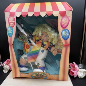 May include: A Cabbage Patch Kids Circus Kid doll with blonde yarn hair, wearing white overalls with green polka dots and a rainbow-striped tie, packaged in a red and white striped box featuring text about a squeaky shoe and a bonus poster.