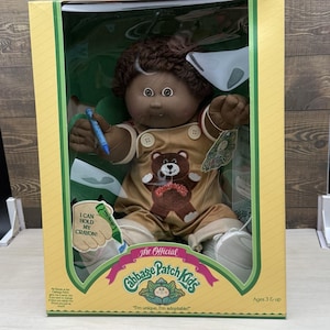 May include: A Cabbage Patch Kids doll with dark brown yarn hair and dark skin, wearing tan overalls with a brown bear graphic. The doll holds a blue crayon. The packaging features the text I CAN HOLD MY CRAYON and The Official Cabbage Patch Kids.