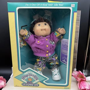 May include: A Cabbage Patch Kids doll in a box with the text I'm A One-Of-A-Kind KID Like You! The doll has dark hair, a purple jacket with yellow buttons, and pants with a black and white zebra print and colorful geometric shapes.