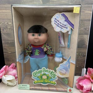 May include: A Cabbage Patch Kids Brushin' Teeth Baby doll in its original packaging. The doll has dark hair, green eyes, and wears a purple and blue outfit with a crocodile graphic. Accessories include a toothbrush, toothpaste, and a bottle.