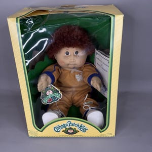 May include: A Cabbage Patch Kids doll with brown yarn hair, freckles, and rosy cheeks, wearing a tan jumpsuit with blue trim and white sneakers. The doll is packaged in a yellow and green cardboard box with the Cabbage Patch Kids logo visible.