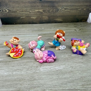 May include: A set of five small cat figurines with brown, pink, and orange hair. One cat holds a red pizza, one is lying down, one is in a blue outfit, one is in a green outfit, and one is in a yellow outfit. Each cat has a small circular tag.
