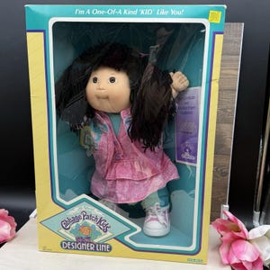 May include: A Cabbage Patch Kids doll in its original packaging, featuring dark brown hair, a pink collared jacket, and white sneakers. The box displays the text I'm A One-Of-A-Kind KID Like You! and includes a birth certificate and adoption papers.