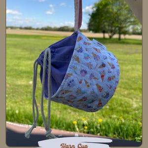 May include: A yarn cup with a light purple interior and a white exterior patterned with colorful ice cream cones. The cup is held by a pink ribbon and has a drawstring closure. The image also shows other yarn cup designs.