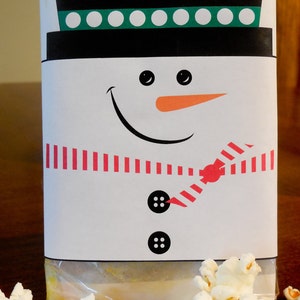 Könnte beinhalten: Ein weißer Schneemann-Geschenkbeutel mit rot-weiß gestreiftem Schal, schwarzen Knöpfen und einer orangefarbenen Karottennase. Der Beutel ist mit Popcorn gefüllt.