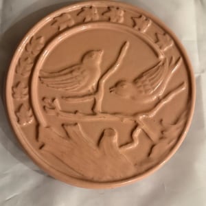 May include: A circular, light peach-colored ceramic mold featuring a relief design of two birds perched on a branch. The border is decorated with a repeating pattern of small, embossed leaves. The surface has a smooth, glossy finish throughout.