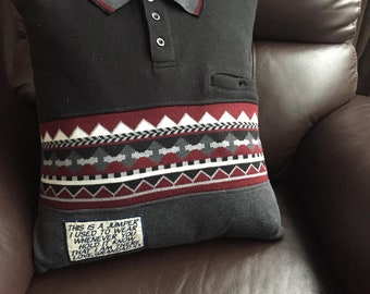 May include: A handmade pillow made from a repurposed black sweater. The pillow features a gray collar with red trim, a patterned band of red, white, and gray, and a small pocket. A label reads, "This is a jumper I used to wear..."