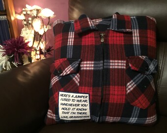 May include: A plaid pillow made from a red, black, and white flannel shirt. The pillow has a zipper and two pockets. A patch with text reads, "HERE'S A JUMPER I USED TO WEAR, WHENEVER YOU HOLD IT KNOW THAT I'M THERE. LOVE, GRANDAD KEN."