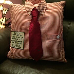 Pode incluir: Uma almofada decorativa projetada para se assemelhar a uma camisa e gravata. A almofada apresenta um padrão xadrez vermelho e branco, uma gravata vermelha e um pequeno bolso. O texto na almofada diz: "THIS IS A SHIRT AND TIE I USED TO WEAR WHENEVER YOU HOLD THEM KNOW THAT I'M THERE. GOD BLESS, GRANDAD XXX."