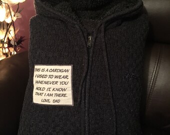 May include: A dark gray hooded cardigan with a zipper closure. A white rectangular patch is sewn on the front, with the text: "THIS IS A CARDIGAN I USED TO WEAR WHENEVER YOU HOLD IT, KNOW THAT I AM THERE. LOVE, DAD."