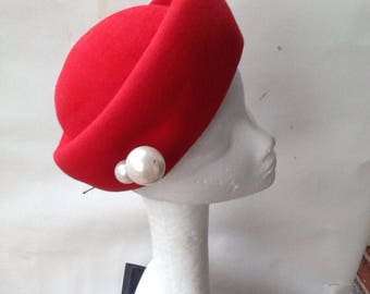 May include: A red felt hat with a rounded crown and a wide brim. The hat is adorned with two large, white pearl-like embellishments.