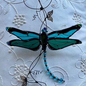 May include: A teal blue stained glass dragonfly with black accents hangs from a wire vine with silver leaves and blue beads.