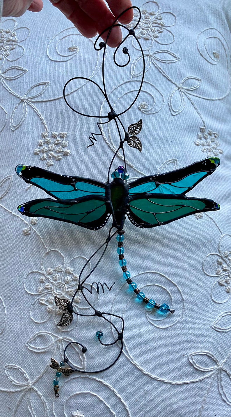 May include: A teal blue stained glass dragonfly with black accents hangs from a wire vine with silver leaves and blue beads.