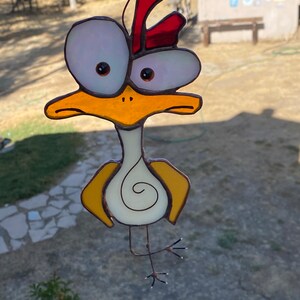 May include: A stained glass chicken with large, white eyes, a red comb, and yellow wings. The chicken is standing on one leg and has a white body with a spiral design.