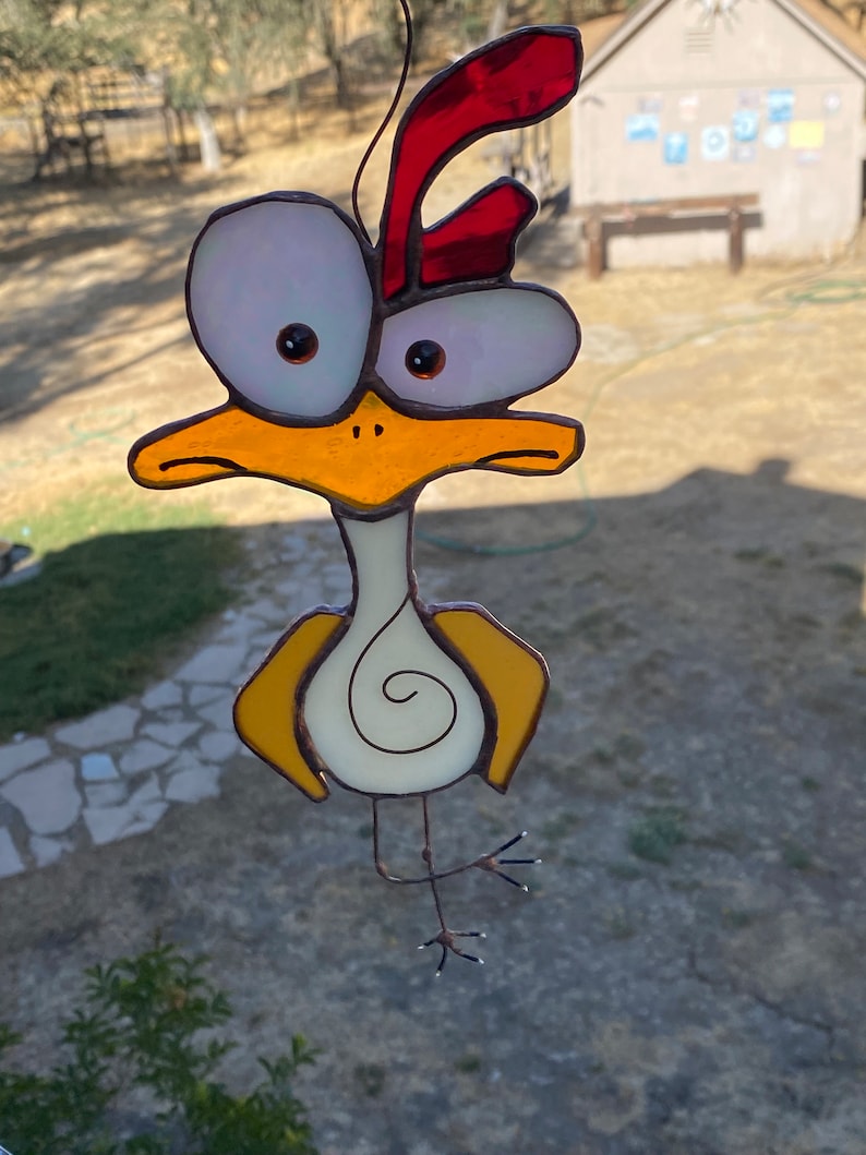 May include: A stained glass chicken with large, white eyes, a red comb, and yellow wings. The chicken is standing on one leg and has a white body with a spiral design.
