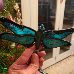May include: A teal blue stained glass dragonfly ornament with black outlining and white dots. The dragonfly has a beaded string hanging from its body.