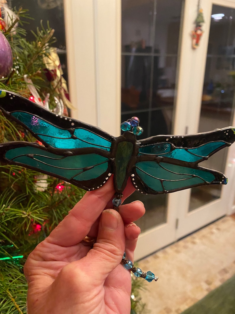 May include: A teal blue stained glass dragonfly ornament with black outlining and white dots. The dragonfly has a beaded string hanging from its body.