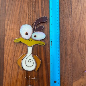 May include: A stained glass bird ornament with brown, yellow, and white glass pieces. The bird has large, round eyes and a long, thin beak. The ornament is hanging from a wire.