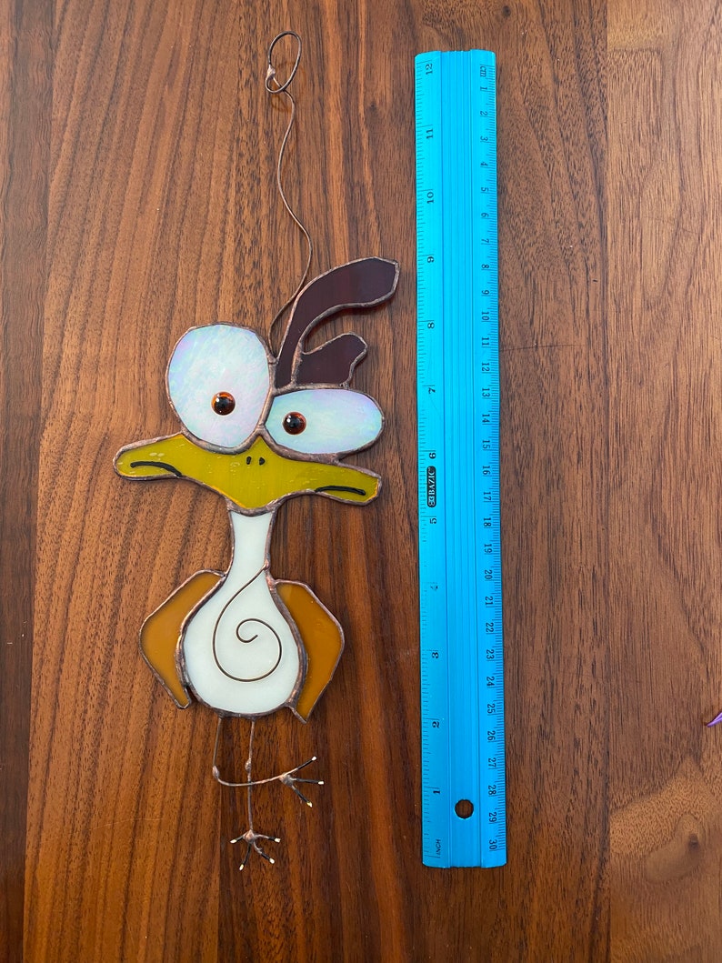 May include: A stained glass bird ornament with brown, yellow, and white glass pieces. The bird has large, round eyes and a long, thin beak. The ornament is hanging from a wire.