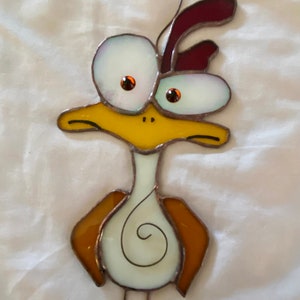 May include: A stained glass bird with large, white eyes, a yellow beak, and brown wings. The bird is hanging from a wire and has a white body with a brown swirl design.