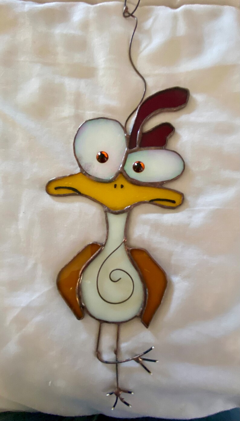 May include: A stained glass bird with large, white eyes, a yellow beak, and brown wings. The bird is hanging from a wire and has a white body with a brown swirl design.