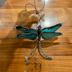 May include: A stained glass dragonfly with teal and green wings hangs from a wire with beads and leaves.