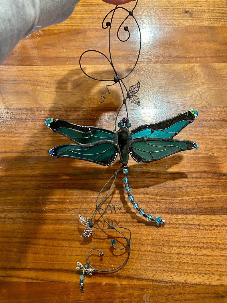 May include: A stained glass dragonfly with teal and green wings hangs from a wire with beads and leaves.