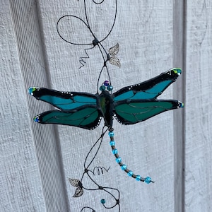 May include: A teal blue stained glass dragonfly with black accents hangs from a wire vine with blue beads and silver leaves.