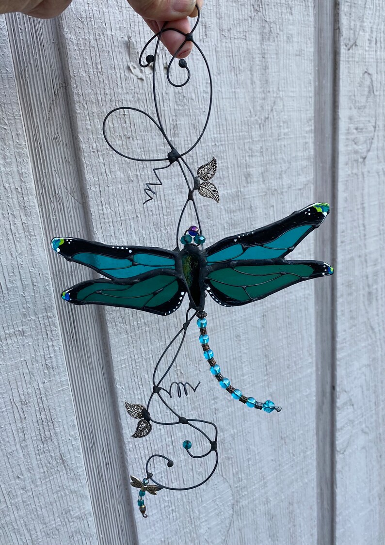 May include: A teal blue stained glass dragonfly with black accents hangs from a wire vine with blue beads and silver leaves.