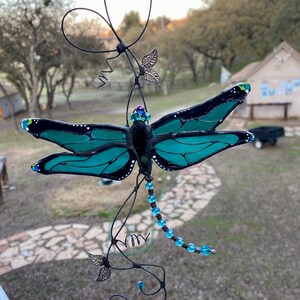May include: A teal blue stained glass dragonfly with black accents hangs from a wire with blue beads and silver leaves.