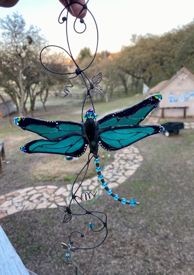 May include: A teal blue stained glass dragonfly with black accents hangs from a wire with blue beads and silver leaves.