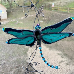 May include: A teal blue stained glass dragonfly with black accents hangs from a wire with silver leaf accents and blue beads.