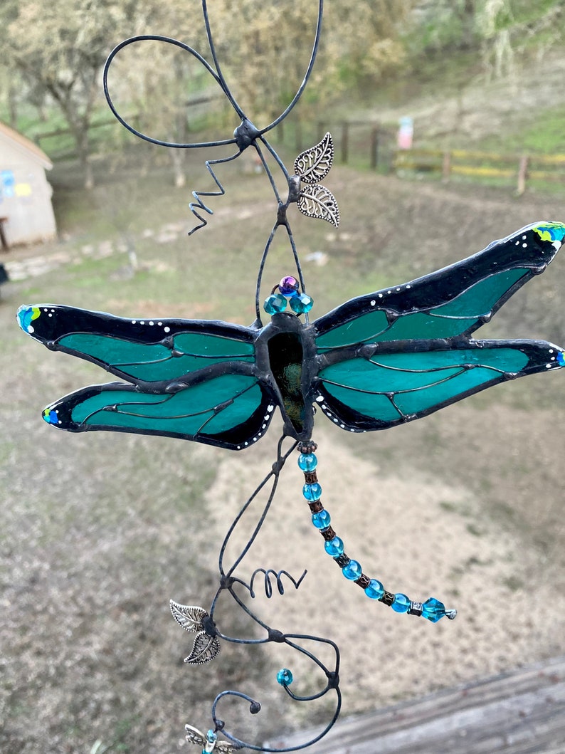 May include: A teal blue stained glass dragonfly with black accents hangs from a wire with silver leaf accents and blue beads.