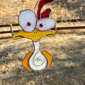May include: A stained glass chicken with large, white eyes, a red comb, and yellow and orange body parts. The chicken is hanging from a wire.