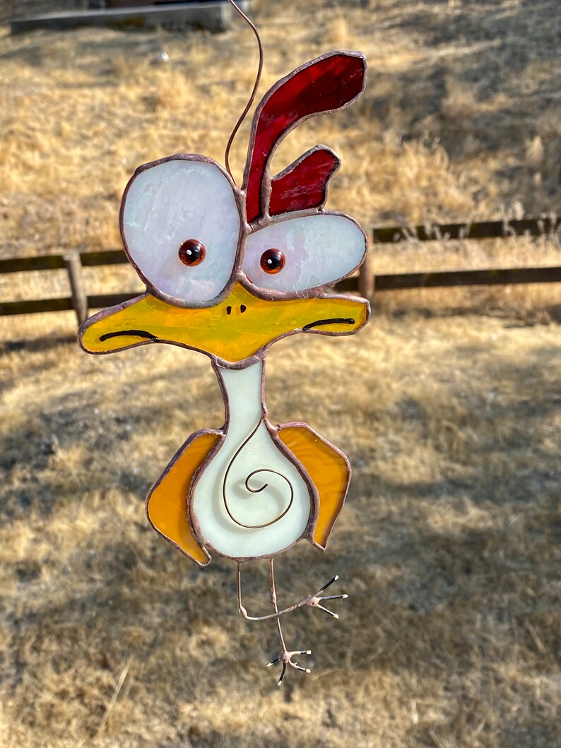 May include: A stained glass chicken with large, white eyes, a red comb, and yellow and orange body parts. The chicken is hanging from a wire.