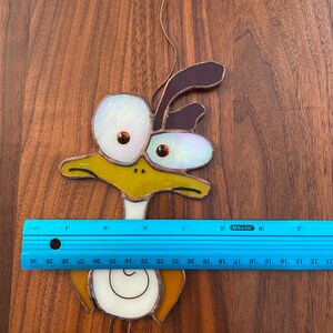 May include: A stained glass bird with large, white eyes and a brown head. The bird is yellow, brown, and white. The bird is hanging from a wire.