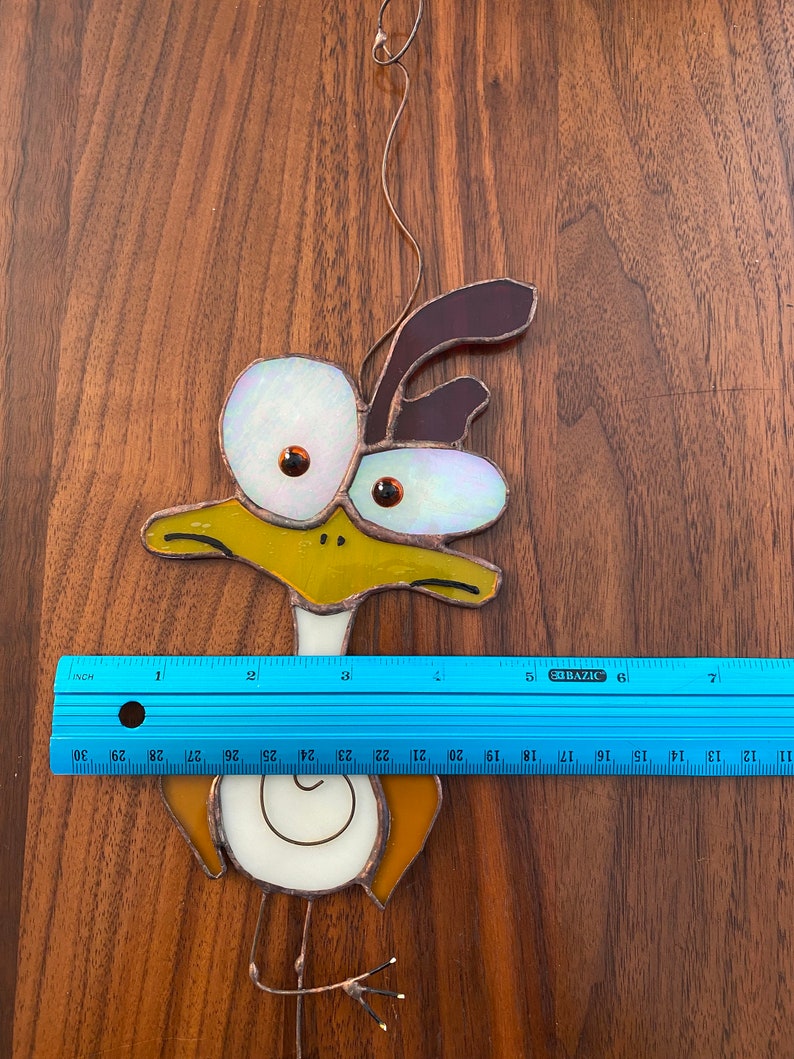 May include: A stained glass bird with large, white eyes and a brown head. The bird is yellow, brown, and white. The bird is hanging from a wire.